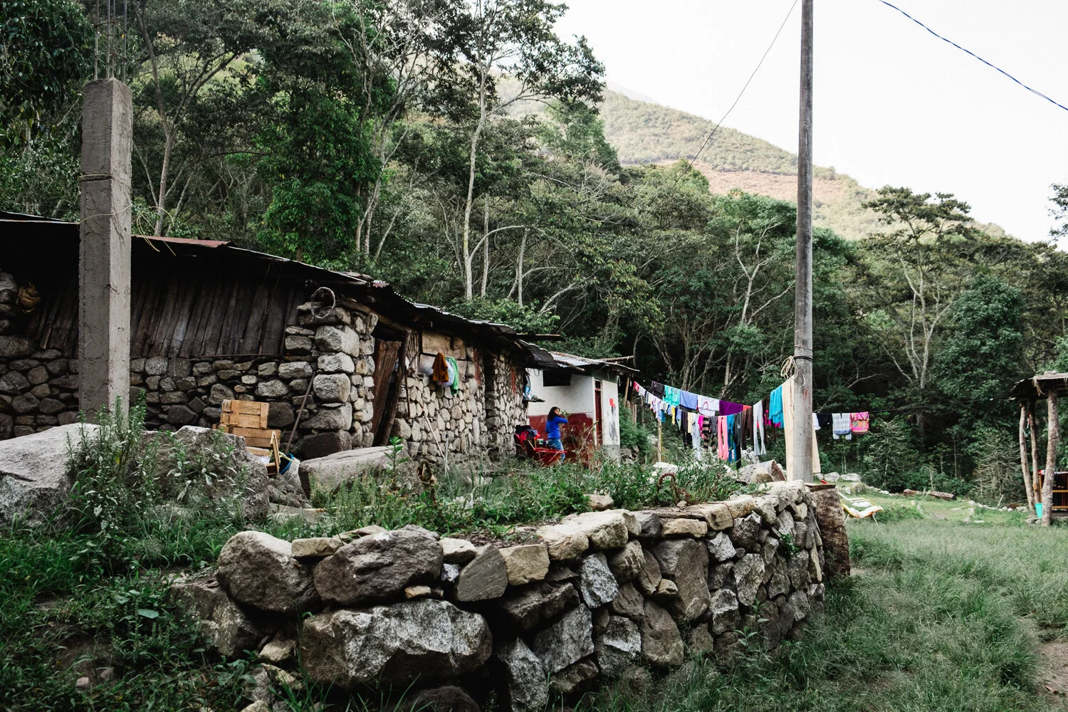 A rural hillside scene with stone and wood houses, colorful laundry hanging on a line, a person sitting outdoors, and a backdrop of a forested mountain.