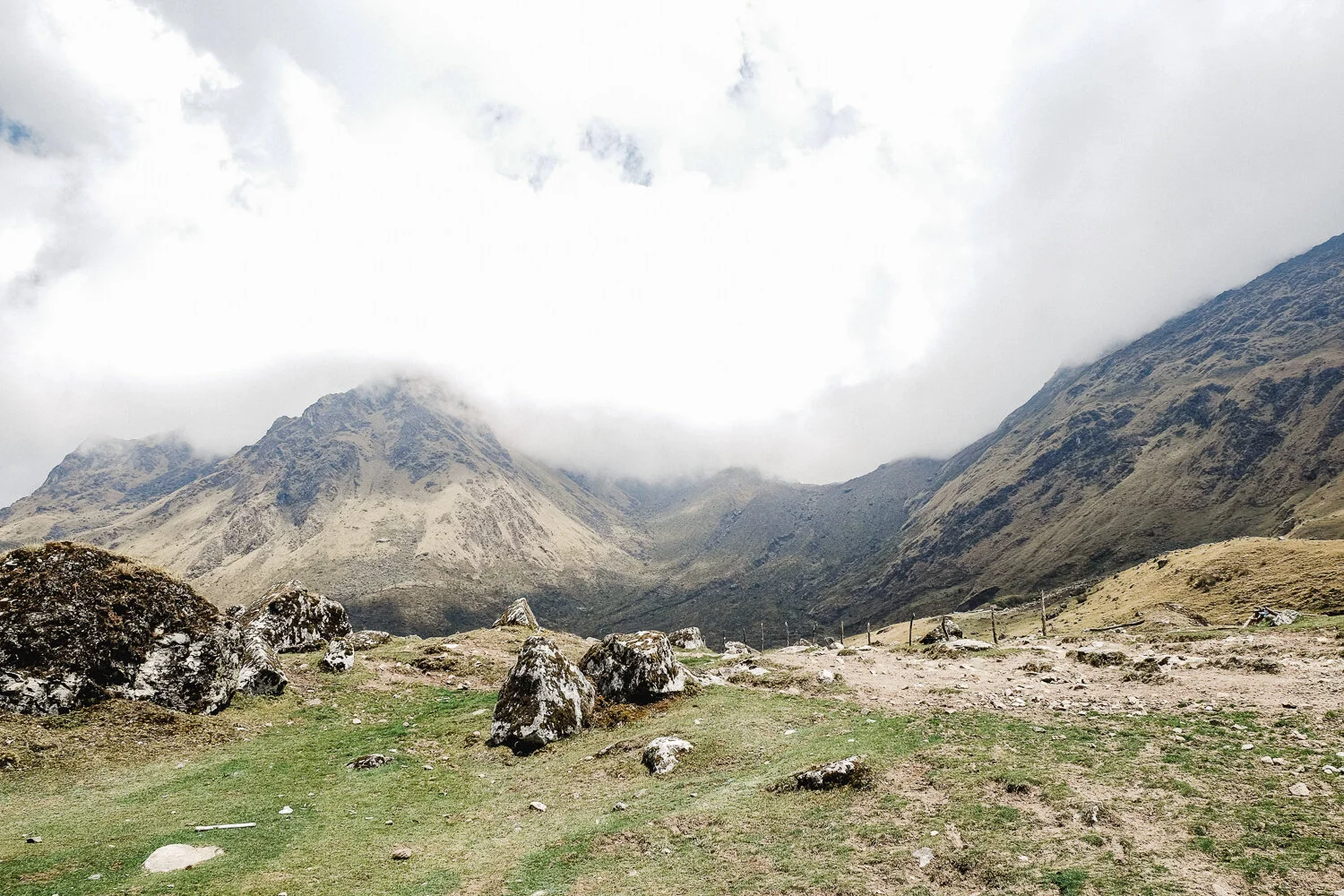 Mountain landscape with rocky foreground, grassy patches, cloudy sky, and mountains partially shrouded in clouds in the background.