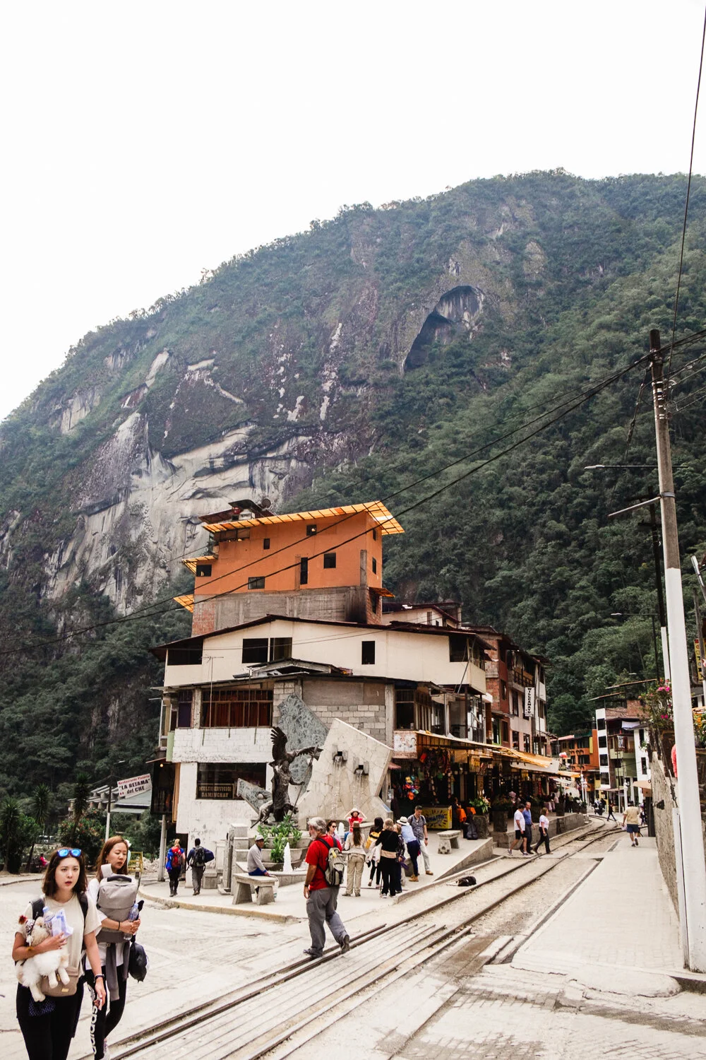 Town with colorful buildings and a mountain backdrop, people walking on the street, train tracks in the foreground, and a statue in the town square.