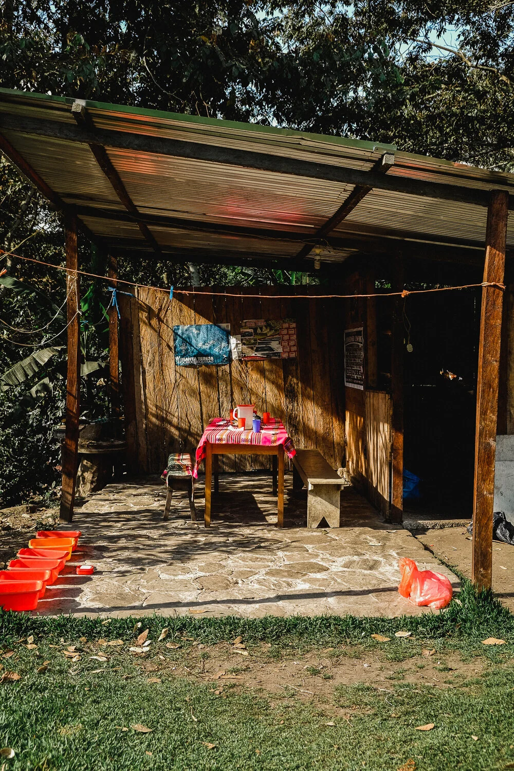 Simple outdoor dining area with a table covered by a red and pink striped cloth, set with cups and bowls, under a metal roof with wooden supports. There are plastic containers on the ground, a bench on one side, and a grassy area in the foreground. G