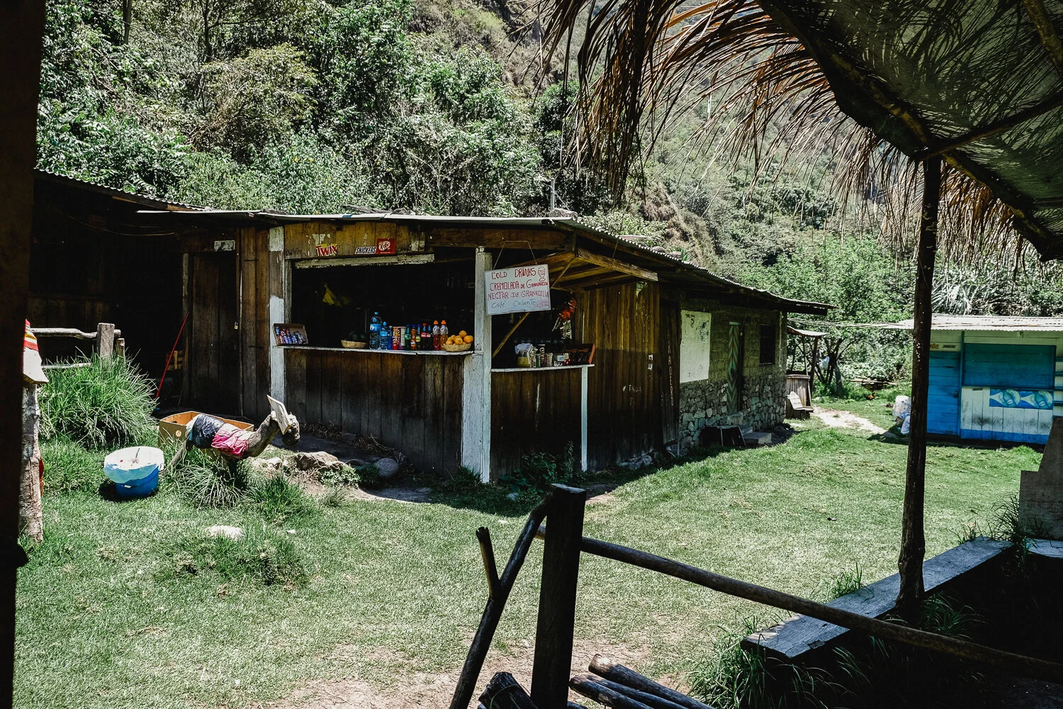 A rustic outdoor village stall with a wooden structure sells drinks and snacks, surrounded by green grass and trees in a rural setting.