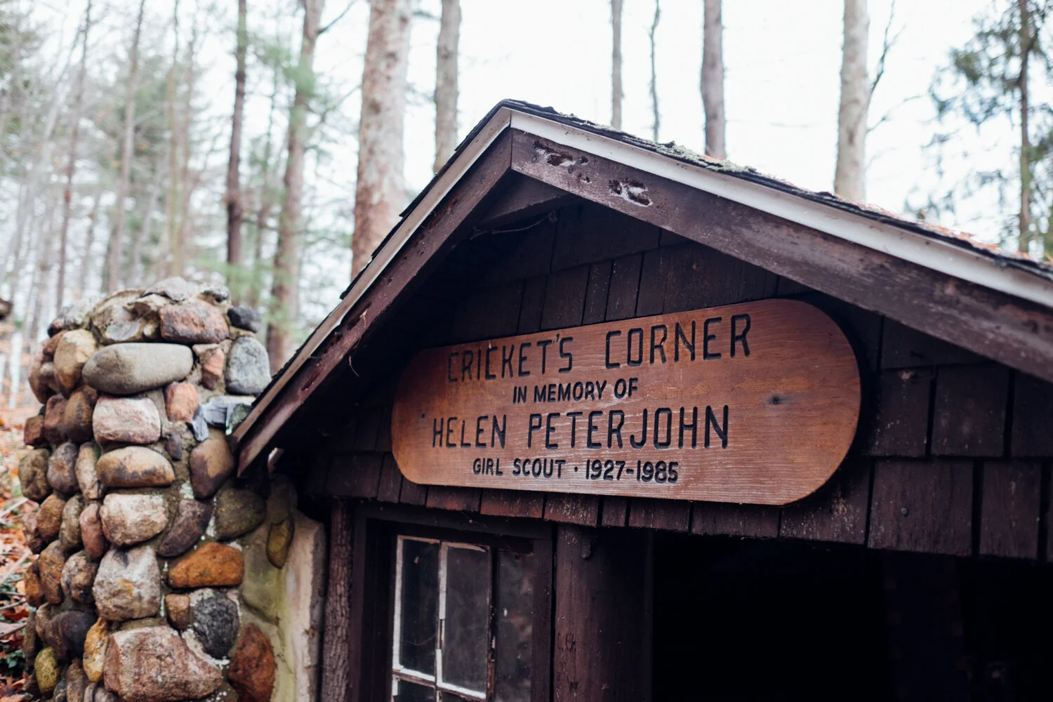 Wooden sign reading 'Cricket's Corner in memory of Helen Peterjohn Girl Scout 1927-1985' mounted on a small wooden shelter with a stone chimney, set in a forest.