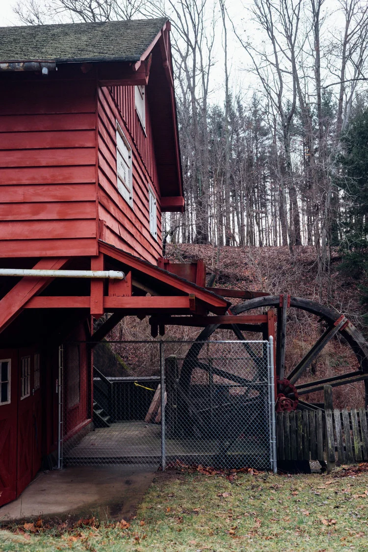 A red wooden watermill with a large wheel, fenced off, set against a wooded hillside in an outdoor rural setting.