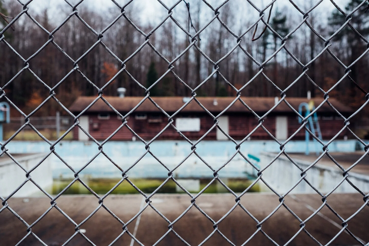 An empty outdoor swimming pool surrounded by a chain-link fence, with a small wooden building and fall trees in the background.