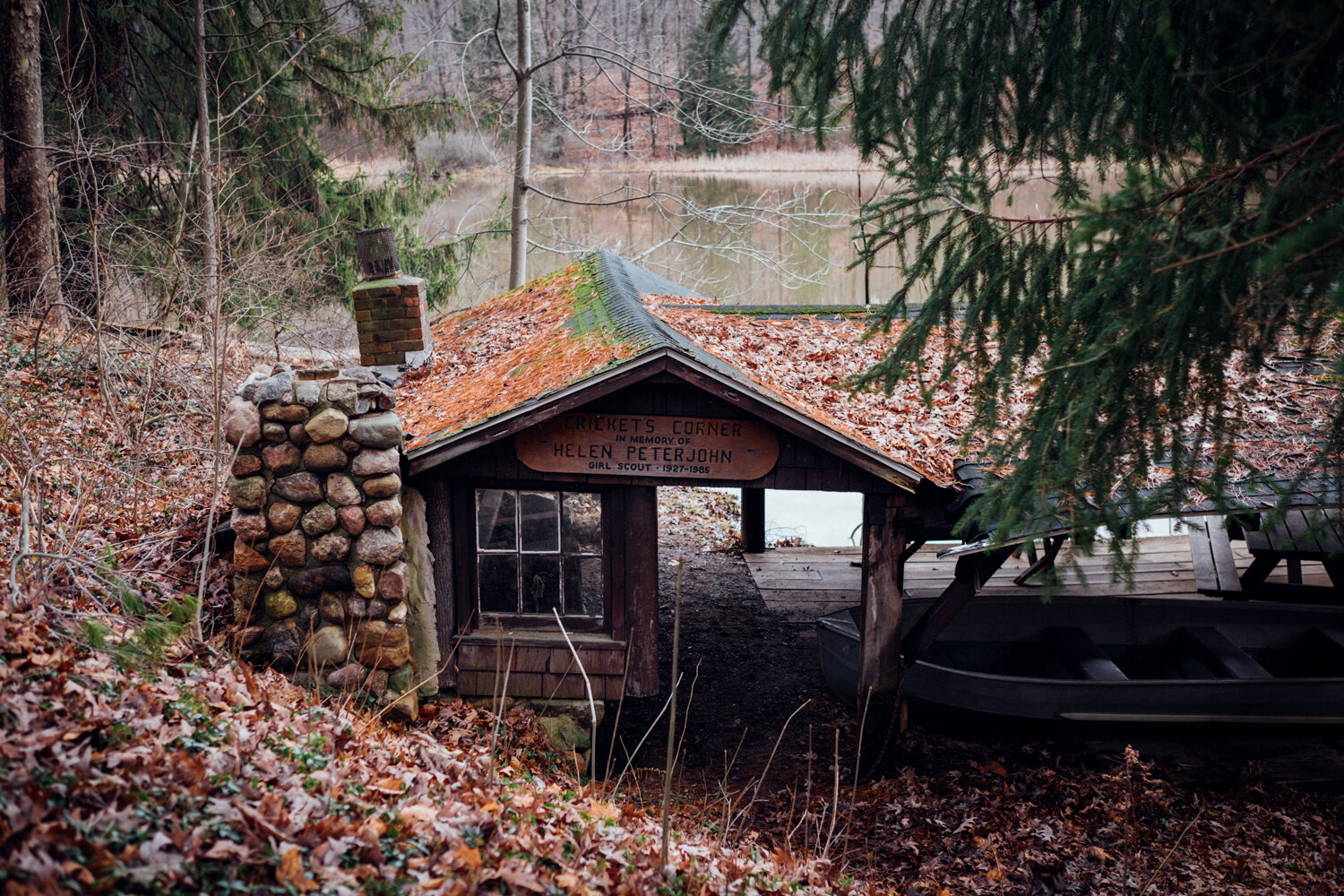A small wooden structure called 'Cricket's Corner' with a stone chimney, surrounded by trees and fallen leaves, next to a body of water in the background.
