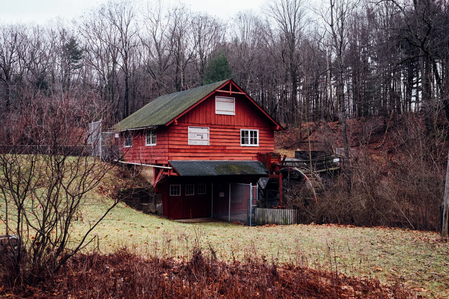 A red old-fashioned watermill house with a sloped roof, positioned on a grassy slope near leafless trees in a wooded area.