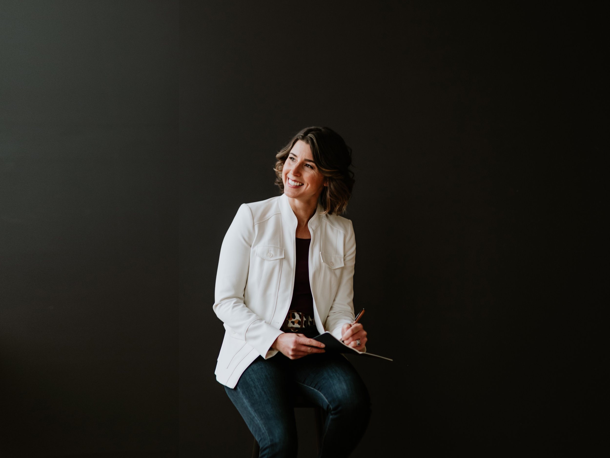 A woman sitting on a stool wearing a white blazer and holding a pen and paper pad smiles as she looks away from the camera