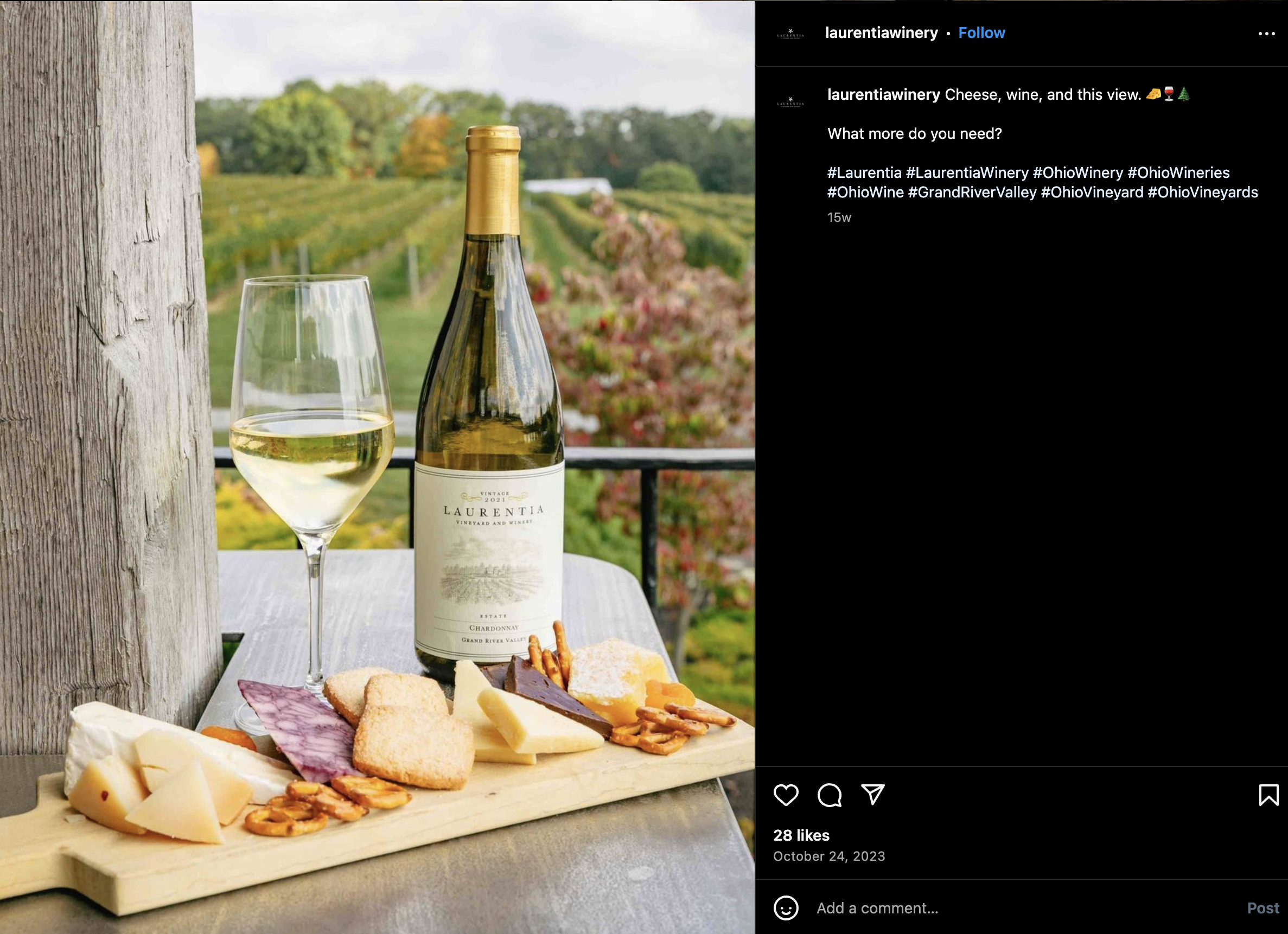 A glass of white wine, a bottle of Laurenti wine, and a cheese and snack platter on a wooden table overlooking a vineyard with colorful fall foliage.