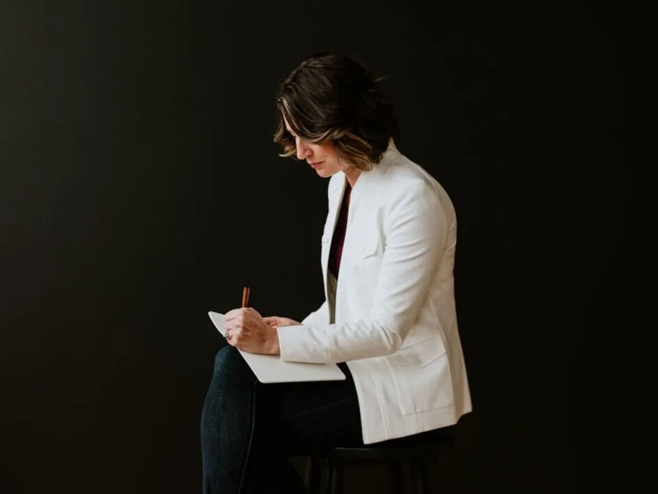Michelle Loufman professional photographer in Cleveland Ohio sits on a chair writing on a notepad in her studio wearing a white blazer