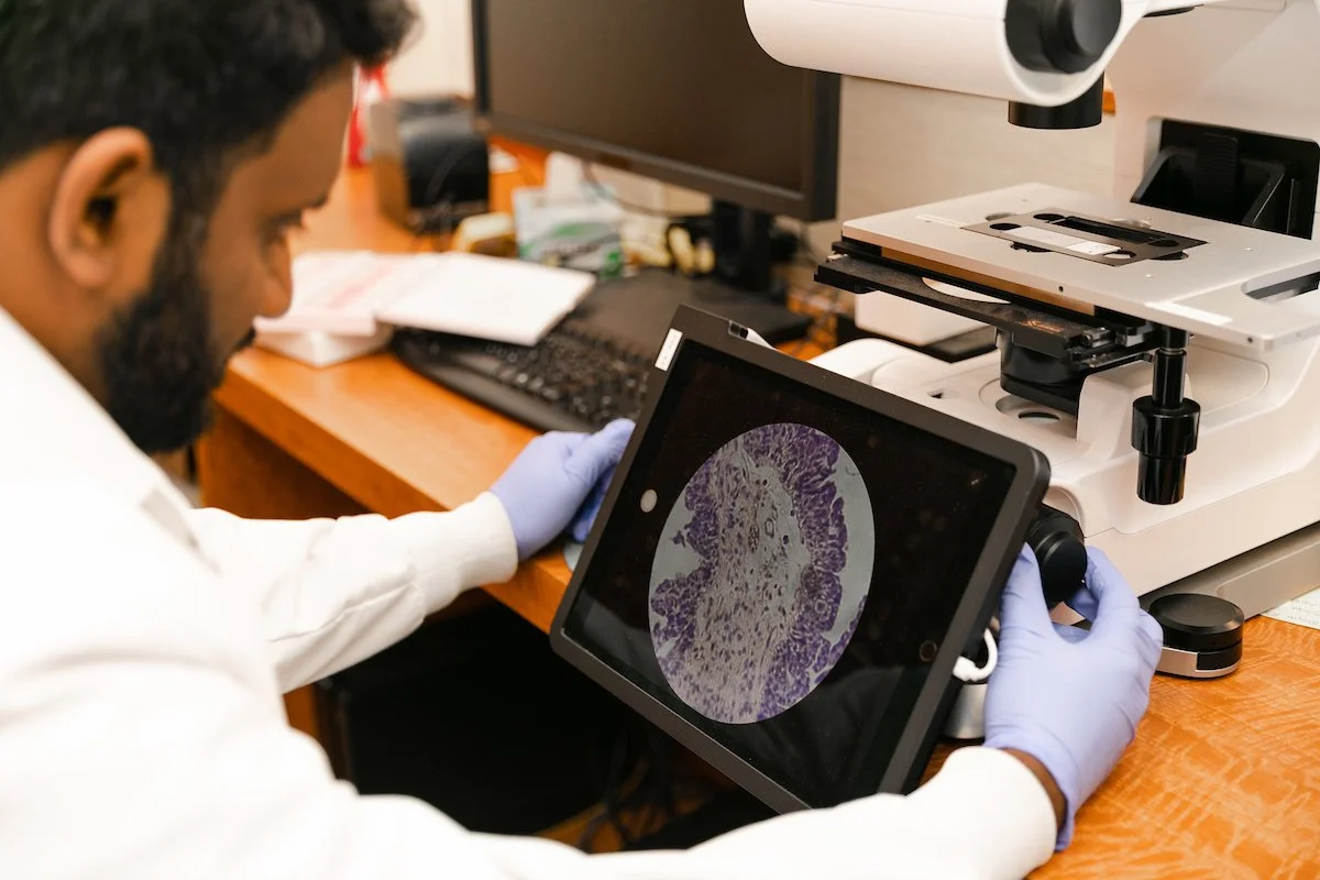 A scientist in a white lab coat and gloves analyzing a magnified tumor cell sample on a tablet connected to a digital microscope at SageMedic.