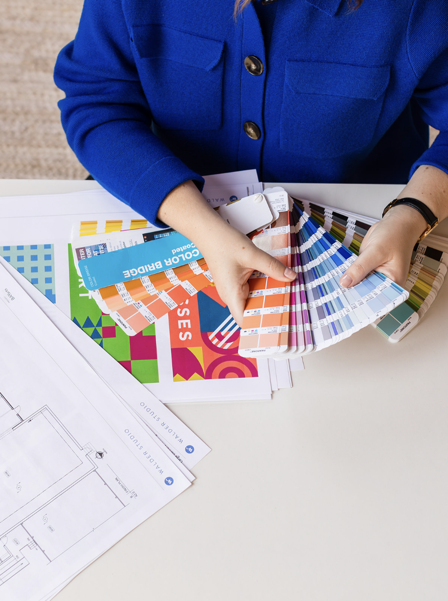 A designer wearing a vibrant blue blazer holds a Pantone color swatch book, reviewing a range of color options. Beneath the swatches, design mockups, architectural blueprints, and branding materials from Walder Studio are spread across the table.