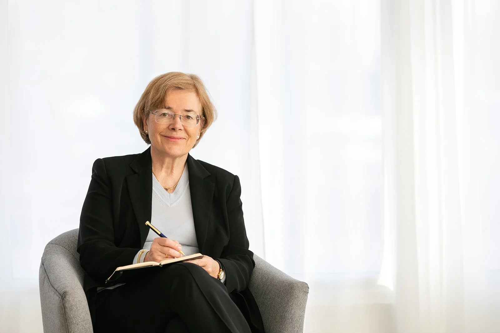 Portrait of psychiatrist Dr. Brigitte Apfel seated in a modern chair, holding a notebook and pen, with soft natural light behind her—professional brand photography for trauma-informed mental health providers.