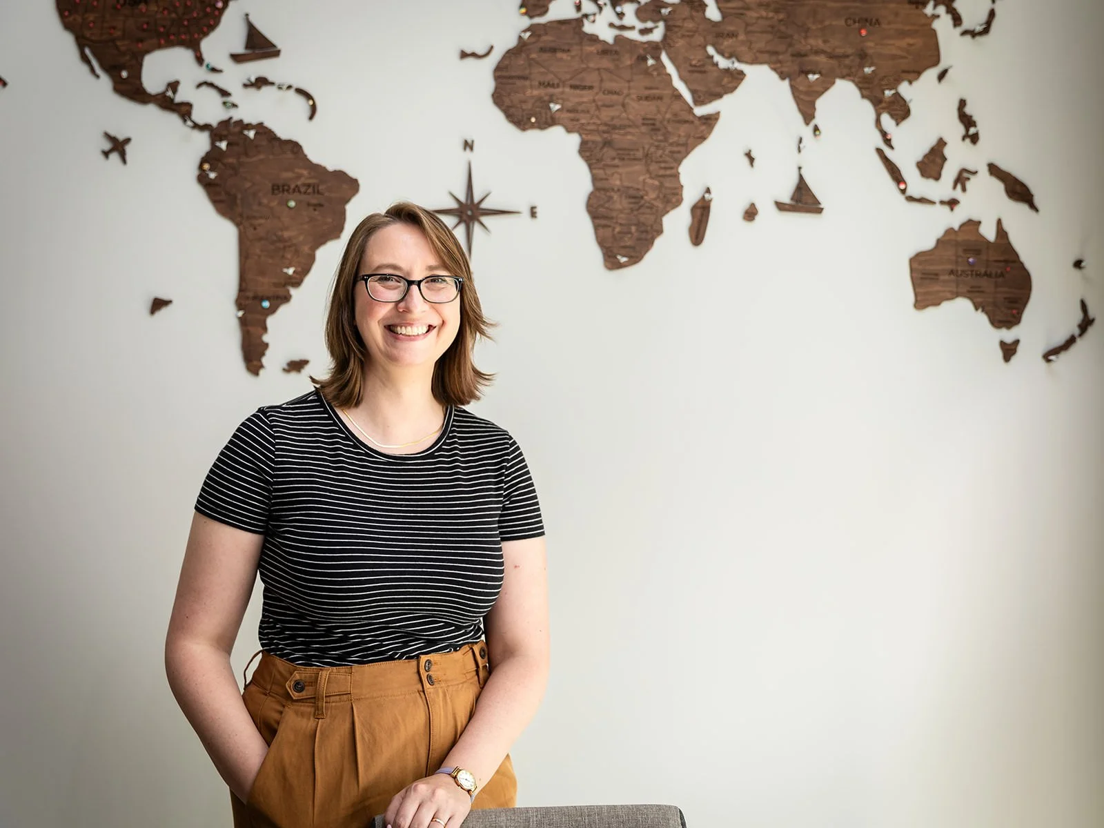 Jessica Gaya standing confidently in front of a wooden world map, smiling, highlighting her role as a travel advisor at Detailed Travel Designs.