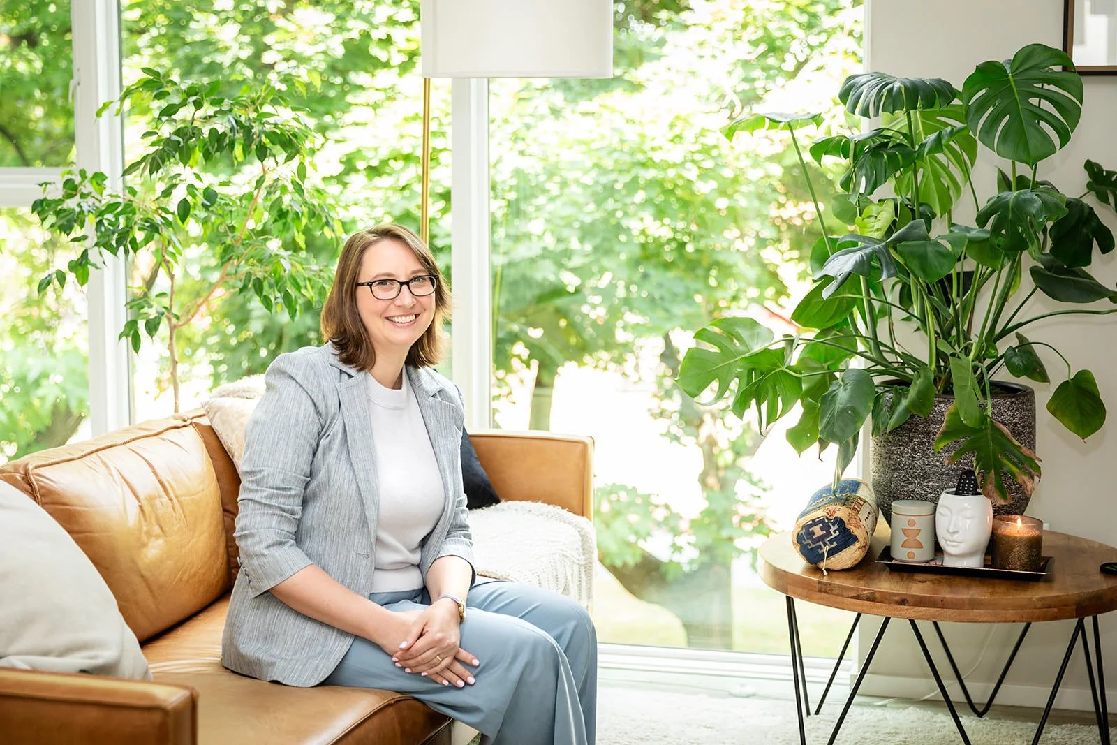 Jessica Gaya sitting on a cozy leather couch in a bright, plant-filled living space, smiling warmly, showcasing her approachable and thoughtful personality as a travel advisor.