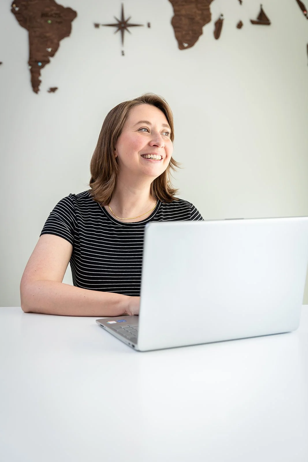 Jessica Gaya sitting at a white desk with a laptop, smiling while working, with a wooden world map behind her, showcasing her dedication to planning personalized travel experiences.