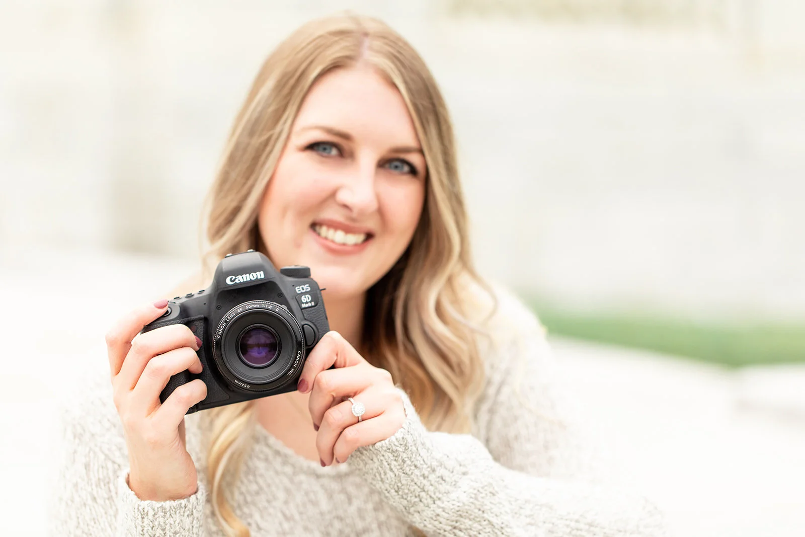 A pretty young woman with blonde hair holds a Canon 6D digital camera and wearing and engagement ring while smiling at the person taking her photo.