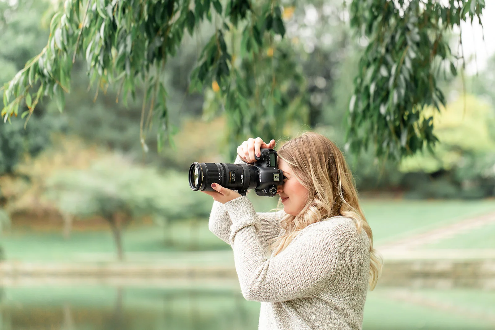 A photographer woman next to a pond surrounded by lush greenery wearing a beige sweater holds a Canon camera.