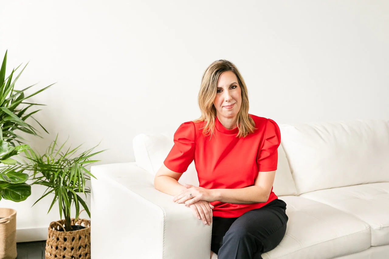 Blonde woman in red blouse posing for headshot on white sofa.