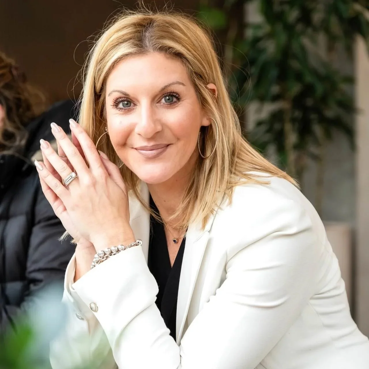 A woman in her 30's with blonde hair wearing a white blazer looks at the camera while her hands touch