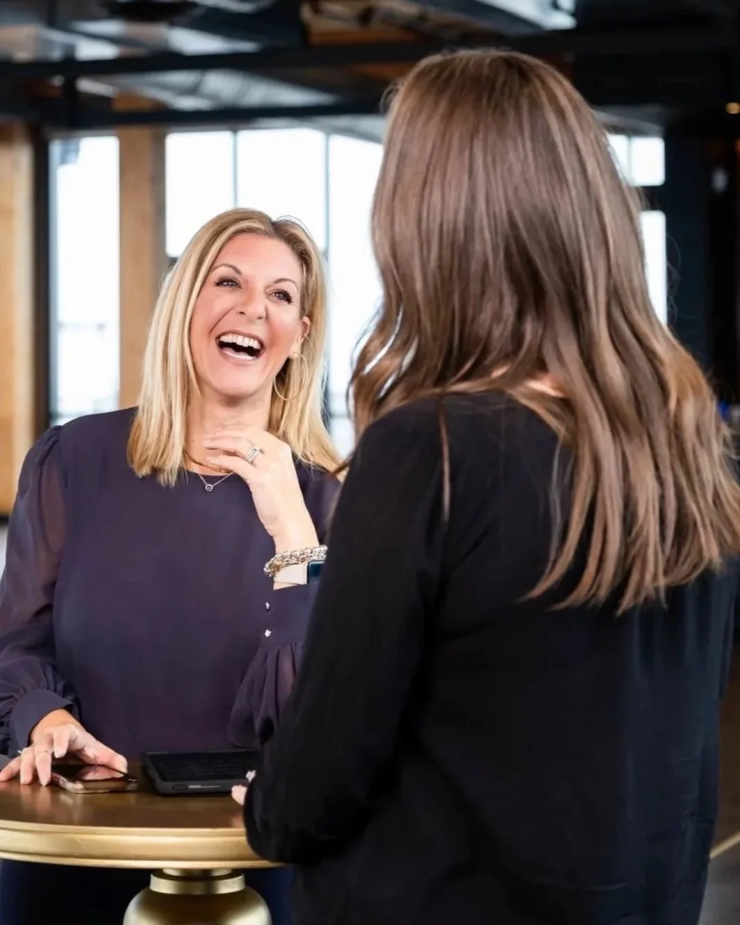 A destination event planner with blonde hair wearing a black shirt clutches her neck and laughs with her client