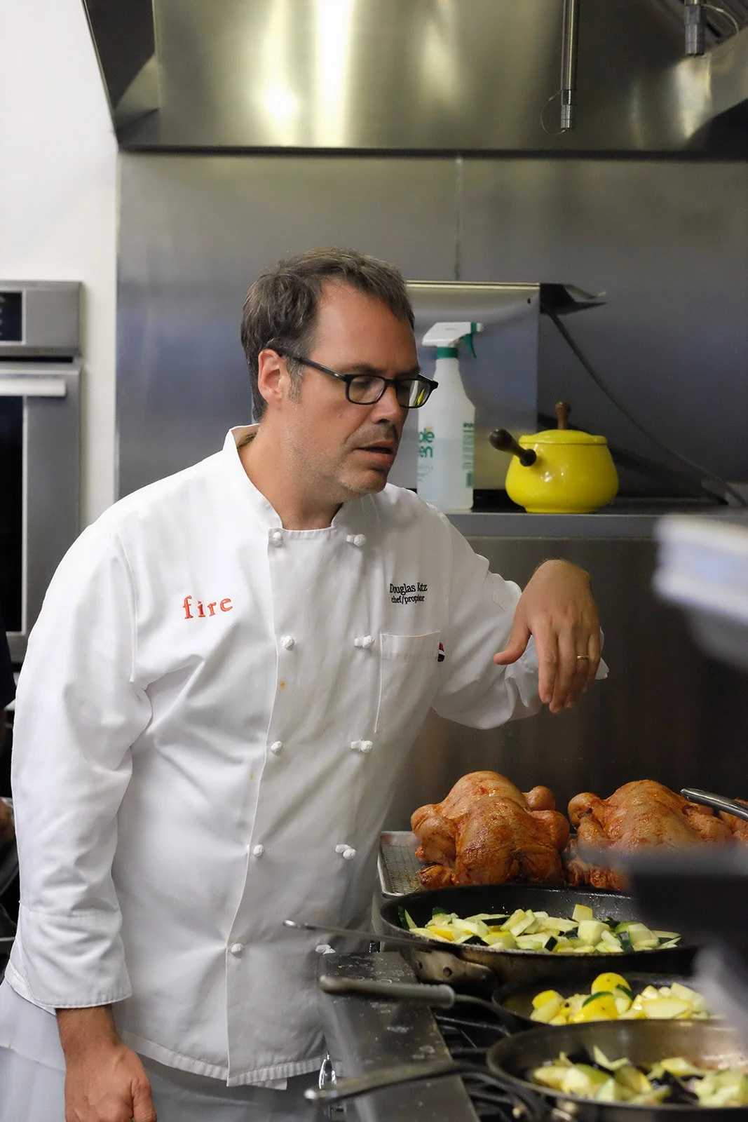 A chef in a white uniform with 'fire' embroidered on the shoulder, and named Douglas Kitz, is in a professional kitchen preparing food, including roasted chickens and chopped vegetables.