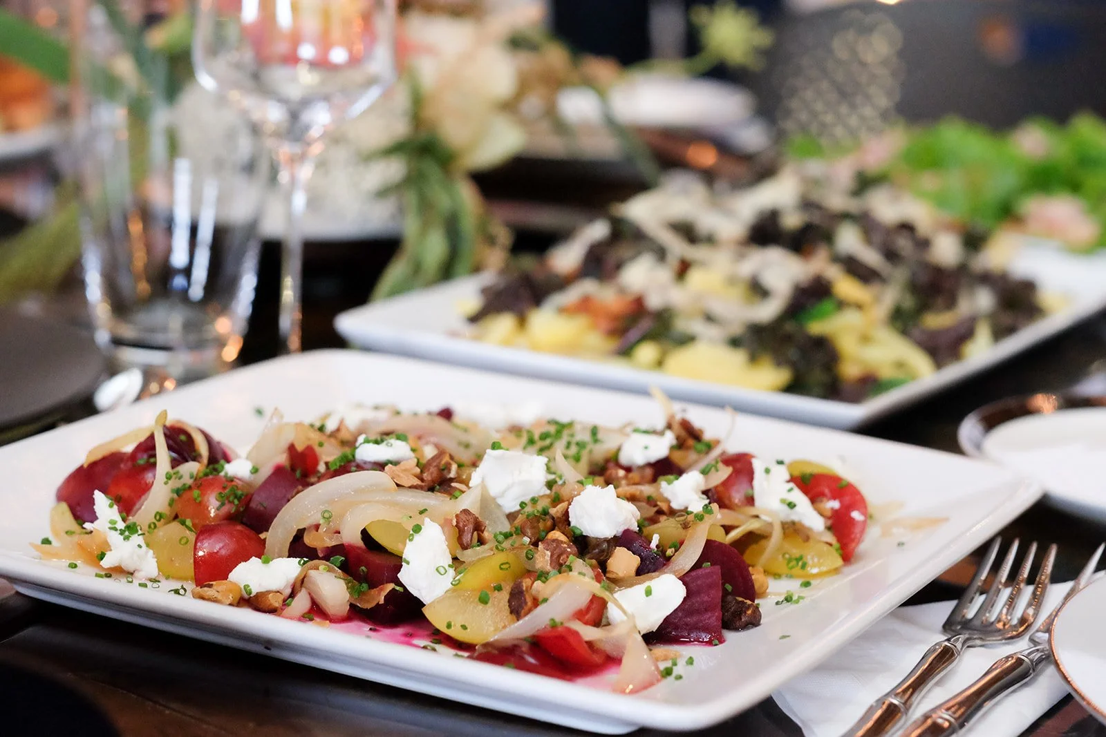 Plate of colorful beet and cherry tomato salad with white cheese, nuts, and herbs, on a table with other dishes, glasses, and cutlery.