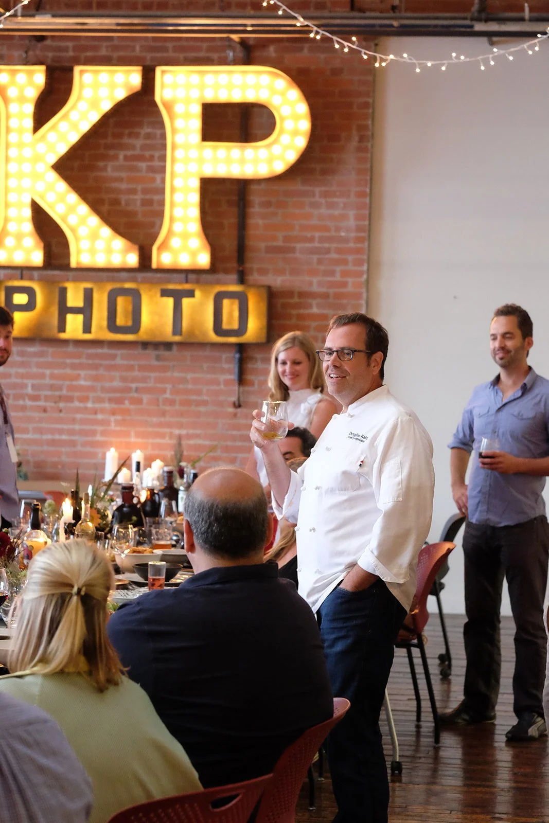 Group of people at a dinner party in a decorated room with a large illuminated sign that reads 'KP PHOTO' on a brick wall. One man, wearing a chef's coat, is standing and holding a drink, smiling, while others sit at a table with candles and decorati