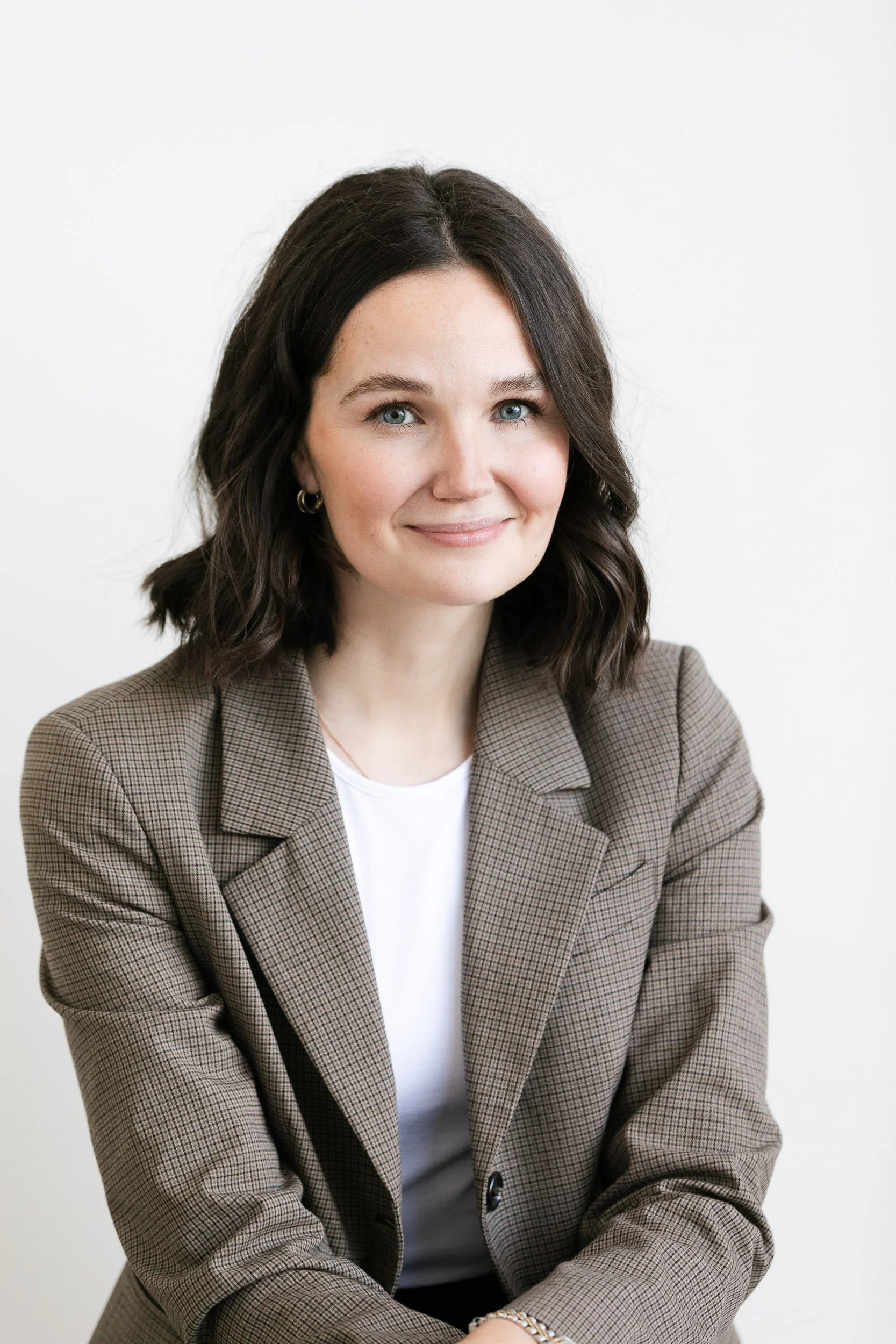 A medical professional with shoulder-length dark wavy hair and blue eyes, wearing a brown checkered blazer over a white shirt, smiling softly, against a plain white background.