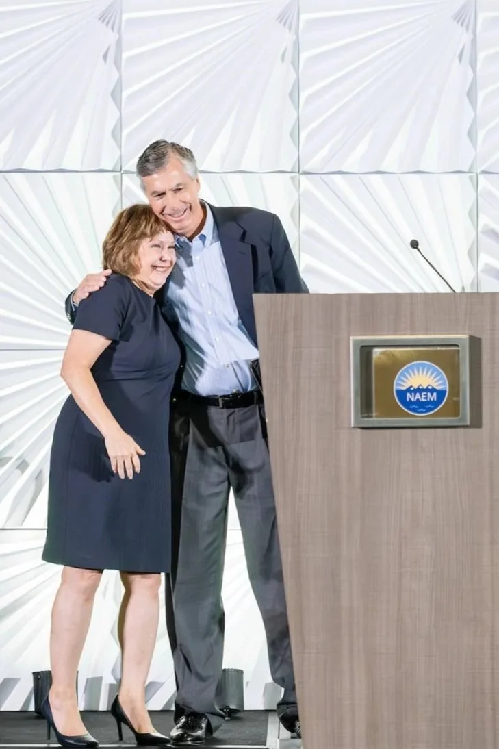 Two people, a woman and a man, smiling and embracing each other at a podium with a NAEM emblem, in a room with a geometric white wall background.