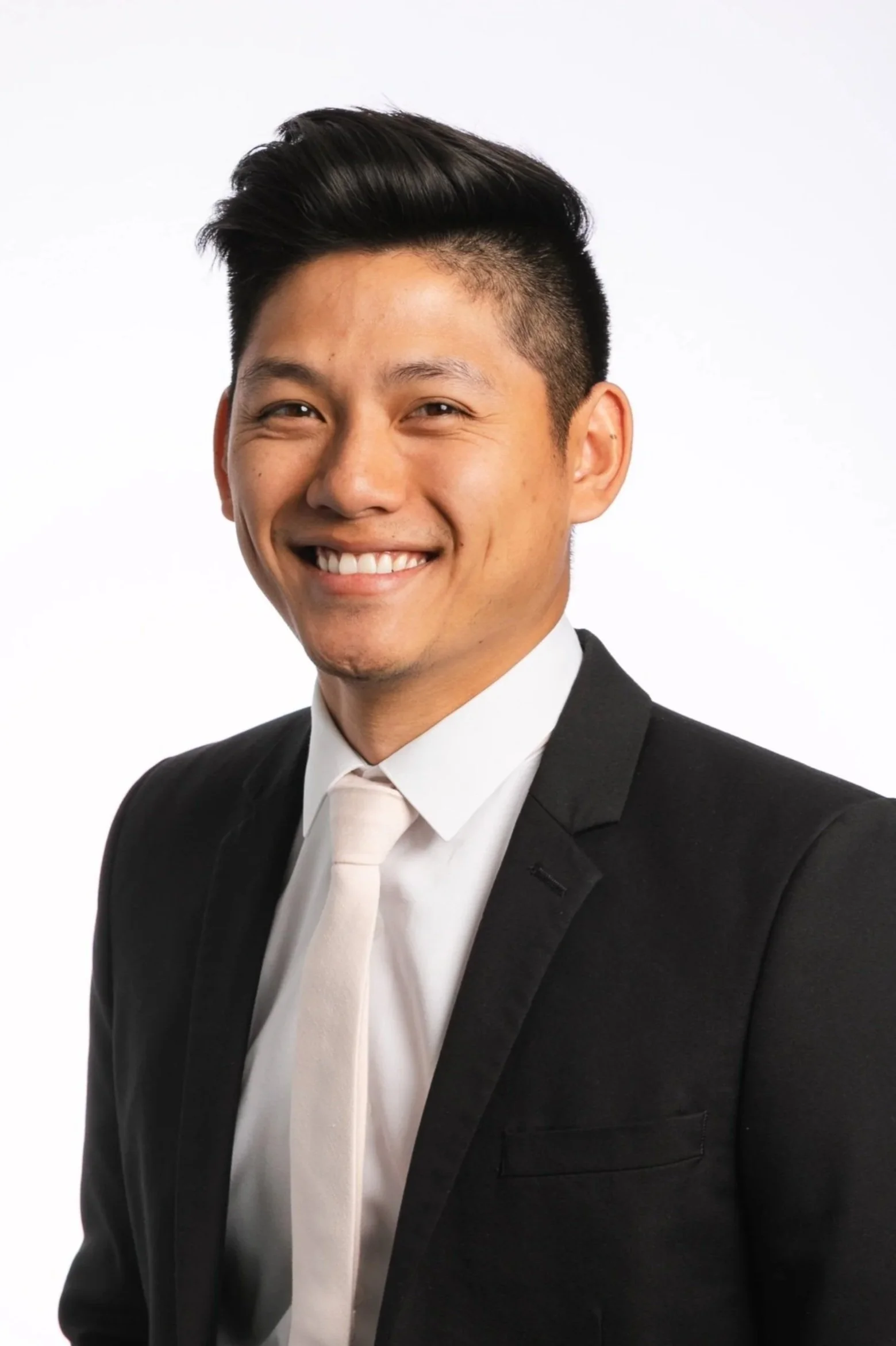 A young man in a black suit, white shirt, and light-colored tie smiling against a plain white background.