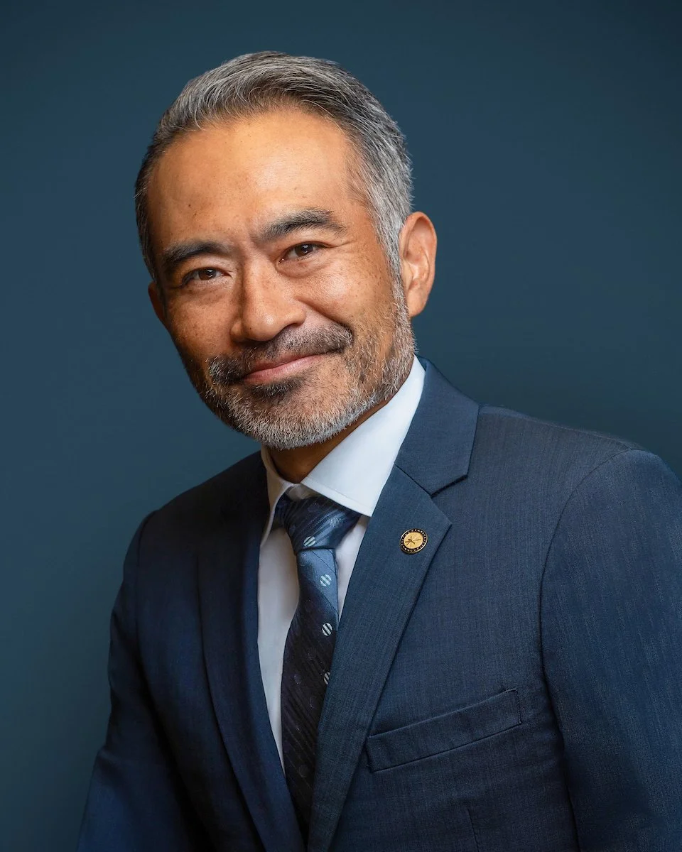 A Korean businessman wearing a blue business suit and a lapel pin, set against a blue backdrop, softly smiles at the camera as a Cleveland photographer captures his executive headshot.