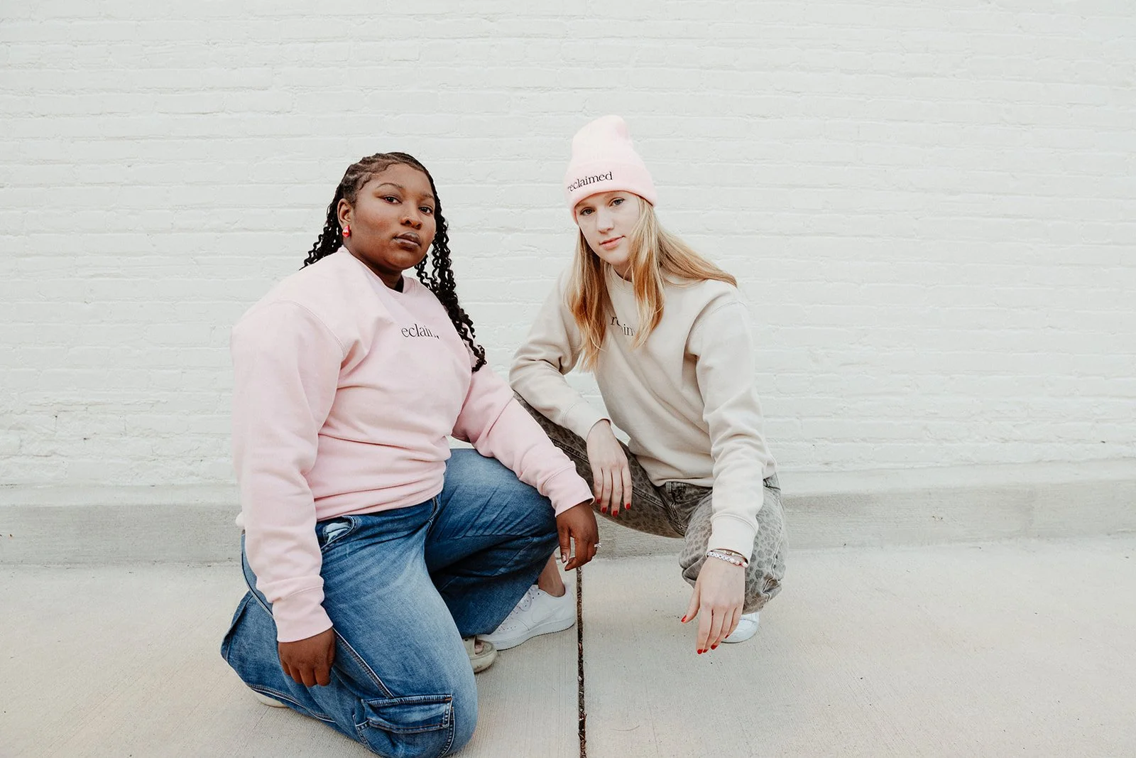 two women squat pose and wearing a pink and beige sweatshirts that say reclaimed