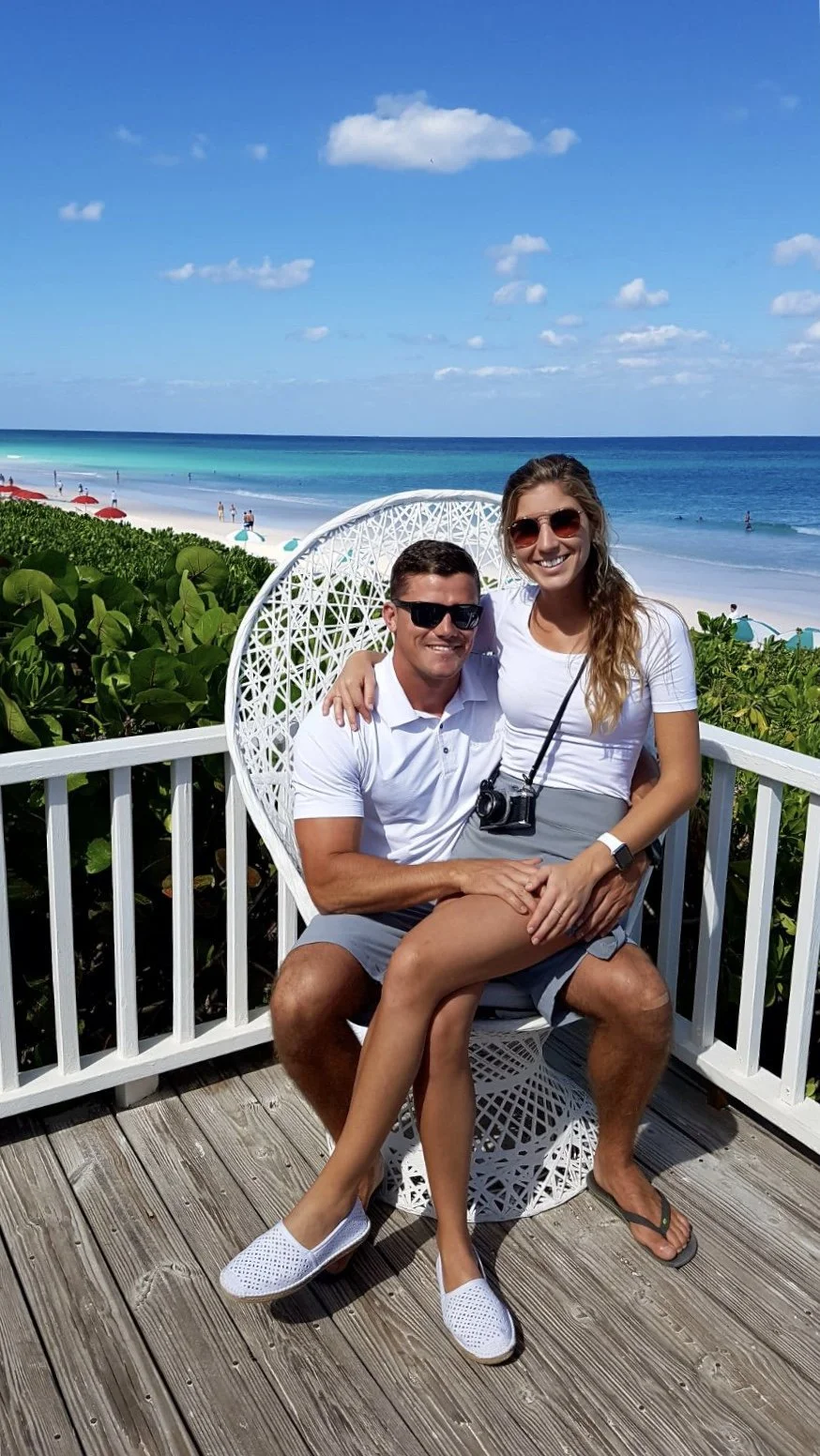 A couple sitting on a white chair on a wooden deck near a beach with blue sky, ocean, and sunny weather. They are smiling, wearing sunglasses, and casual summer clothes.