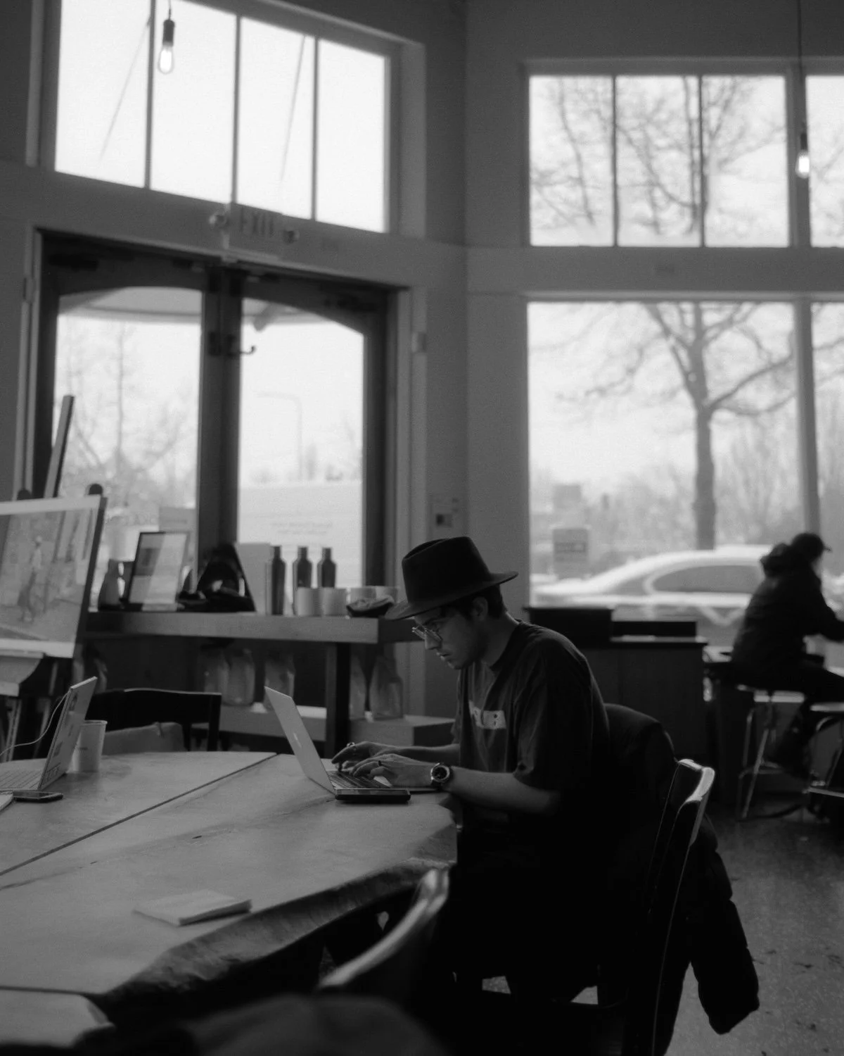 A man wearing a hat and glasses working on a laptop in a cafe with large windows and another person seated in the background.