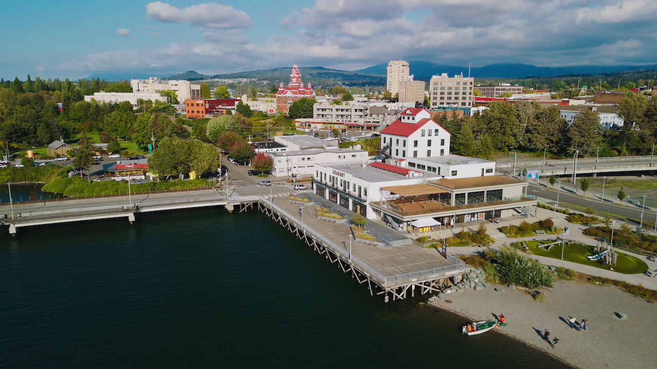 Aerial view of a waterfront town with a pier, colorful buildings, a park, and a mix of trees and mountains in the background under a partly cloudy sky.