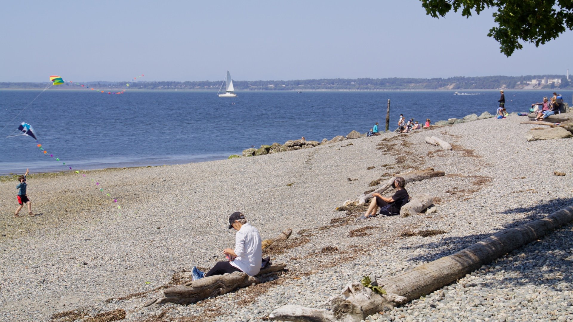 People relaxing and flying kites on a pebble beach near the water. Sailboats are seen in the distance on a sunny day with a clear sky.