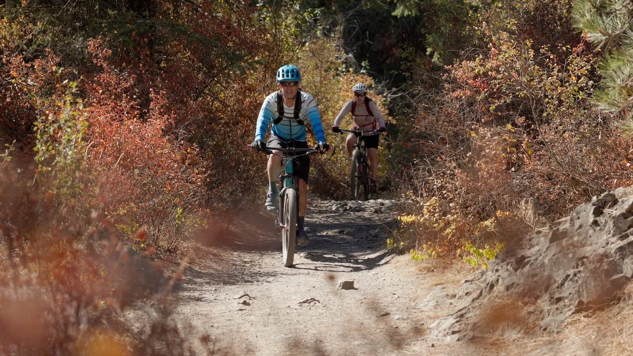 Two women riding mountain bikes on a wooded trail surrounded by autumn foliage.