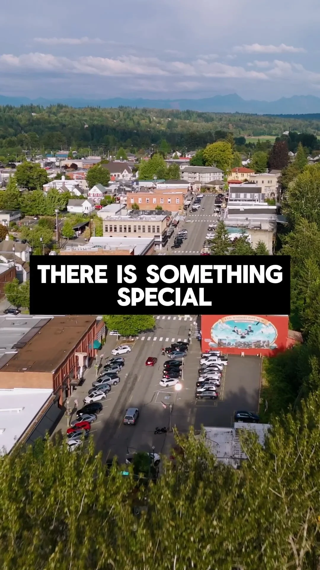Aerial view of a small town with a mixture of commercial and residential buildings, parking lots, and surrounding trees, with mountains in the background.