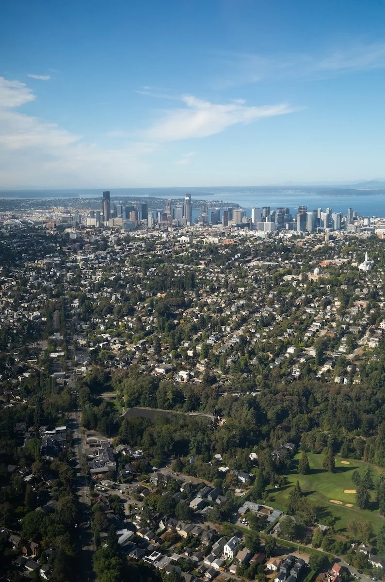 Aerial view of a city skyline with tall buildings along the waterfront, with a residential neighborhood, green parks, and tree-lined streets in the foreground under a blue sky.