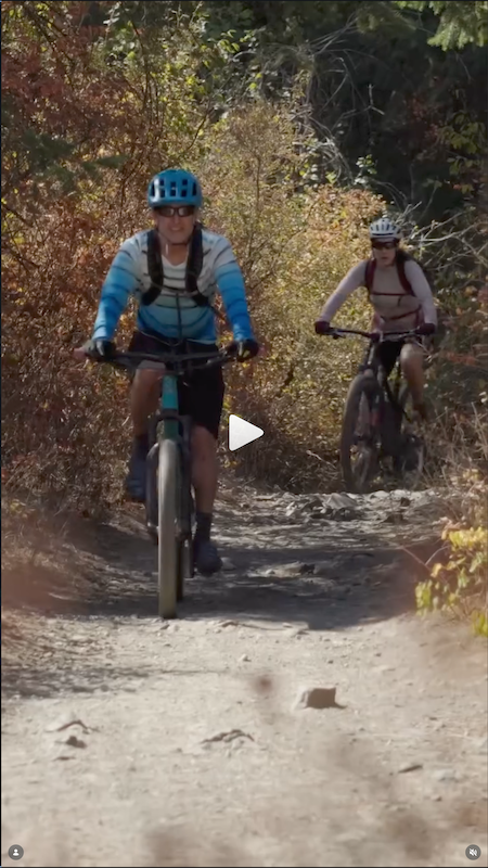 Two women mountain biking on a dirt trail surrounded by trees and foliage.