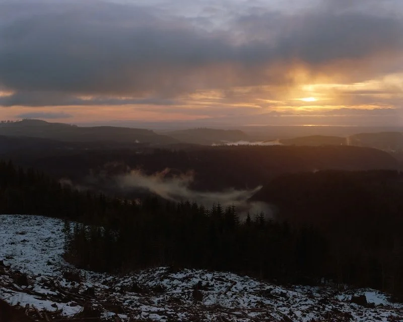 Sunset over a mountainous landscape with a forested valley and patches of snow on the ground.