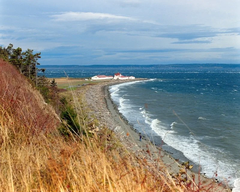 A coastline with a grassy hill on the leftside, a pebble beach, and a distant building on the shoreline, under a cloudy sky.