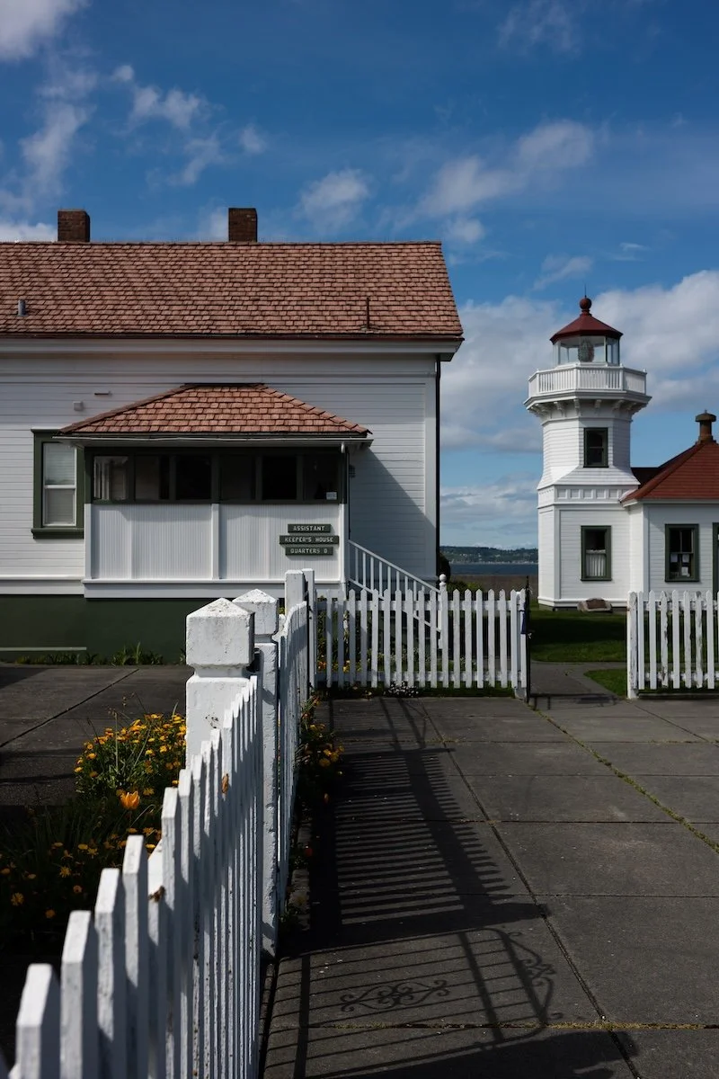 A white house with a red tile roof and a small porch, near a white lighthouse with a red top. A white picket fence surrounds the porch area, with a sidewalk and yellow flowers nearby. The sky is partly cloudy.