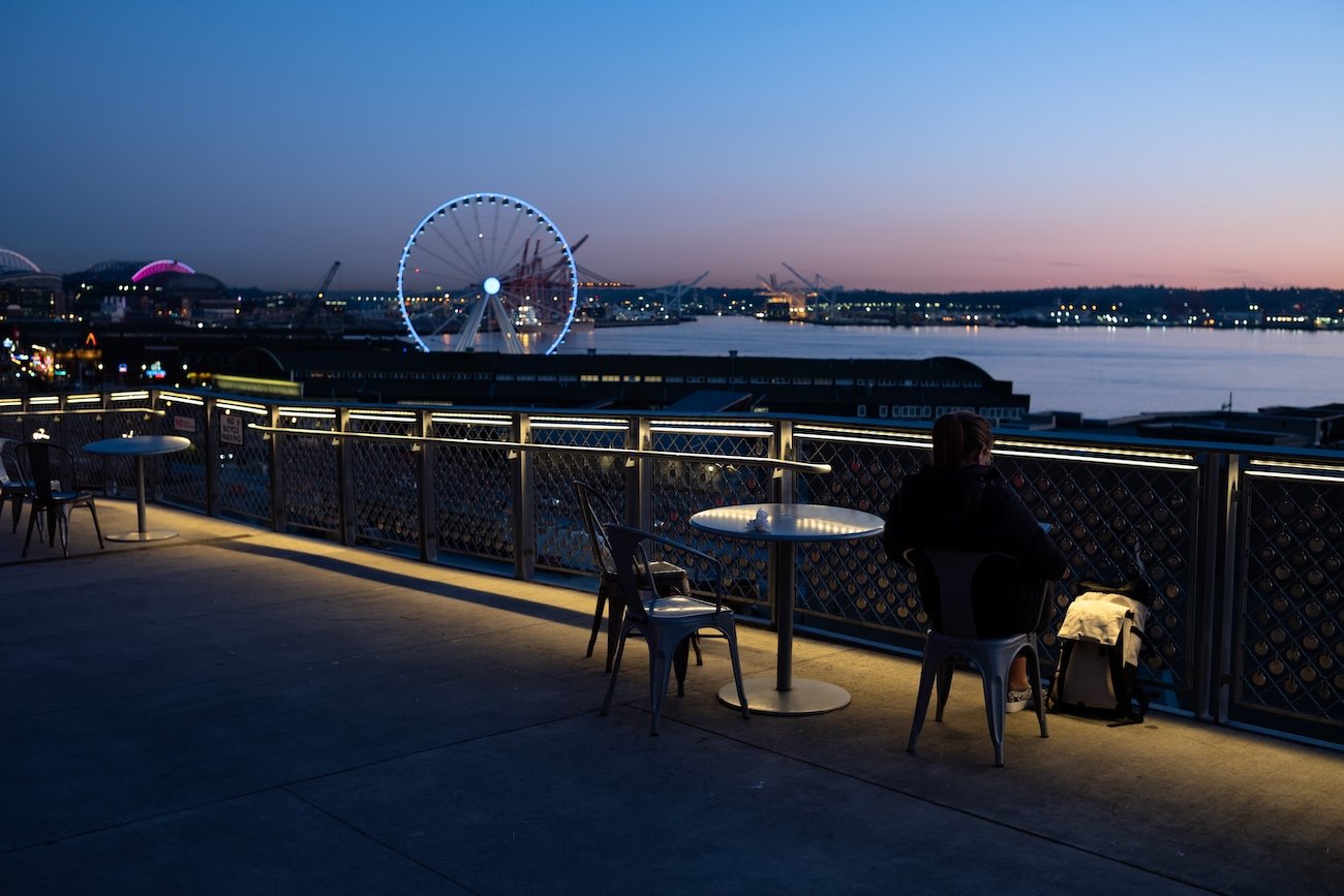 A person sitting alone at a table on a waterfront balcony during evening, overlooking a city skyline with a lit Ferris wheel and bridge across the river.