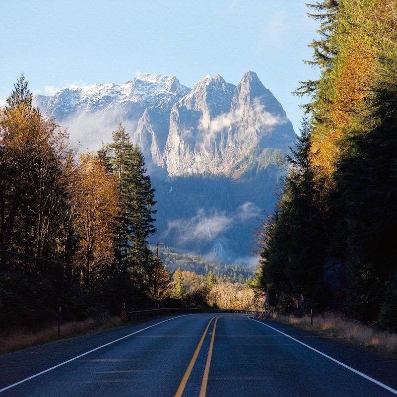 A two-lane road with double yellow lines, flanked by trees, leading towards a mountain range with snow-capped peaks in the distance under a clear blue sky.