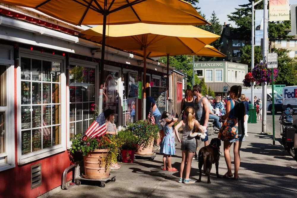 People standing in line outside a bakery or shop with yellow umbrellas and potted flowers on a sunny day.
