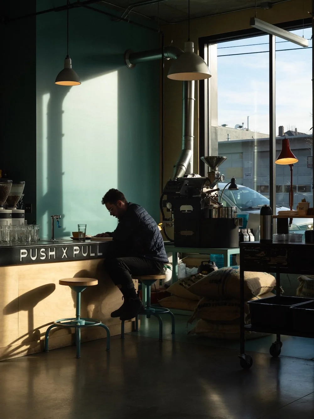 Interior of a coffee shop with sunlight streaming through large windows, a barista preparing drinks, and seating area with stools and hanging lamps.