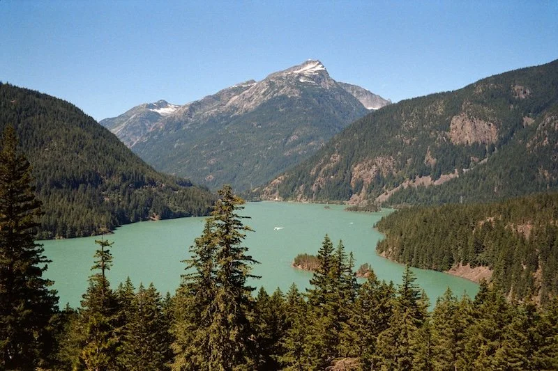 A lake surrounded by green forested mountains with snowy peaks in the background under a clear blue sky.
