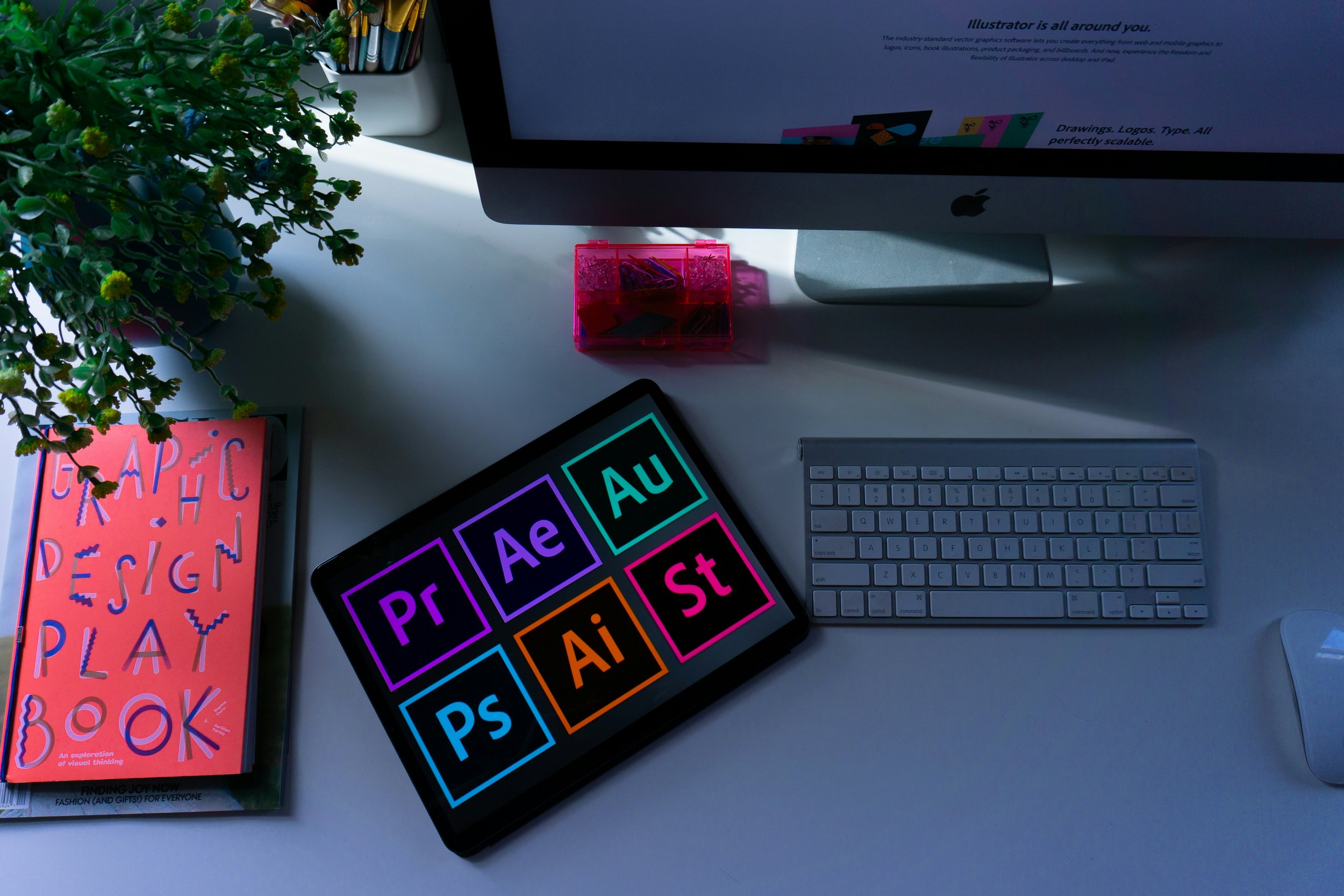 Desk with a large monitor, a keyboard, a mouse, a tablet with colorful Adobe software icons, a closed pink design book, a plant, and a container of colored pens.
