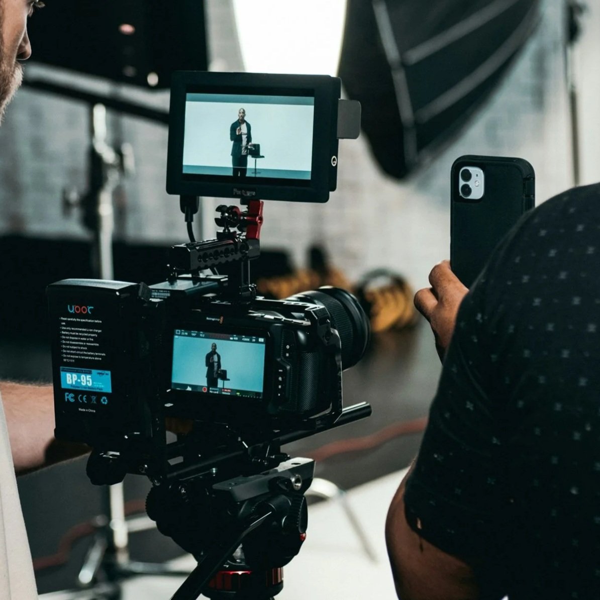 A person filming a presentation with a professional camera, while another individual watches and takes a photo using a smartphone. The camera's monitor shows the presenter standing at a small table with equipment.